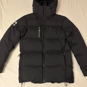 Svart dunjacka Sail Racing med huva - Svart dunjacka från Sail Racing med huva och dragkedja framtill. Jackan har logga på bröstet och ärmen, samt flera praktiska fickor. Puffig design med breda kanaler och justerbar nederkant. Perfekt för kalla dagar och riktigt snygg streetstil. Användning minimal. Kvitto finns.