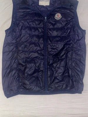 Moncler väst - Snygg mörkblå dunväst från Moncler med klassisk quiltad design och dragkedja framtill. Västen har Moncler-logga på bröstet och är tillverkad i glansigt material som ger en sportig look. Perfekt för lager-på-lager-stil under kyliga dagar. Dragkedjan är dock sönder längst ner så den skulle behöva bytas, annars måste man ta på sig den som en hoodie