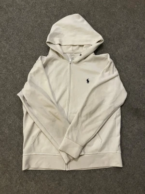 Vit hoodie från Polo Ralph Lauren - Snygg vit hoodie med dragkedja från Polo Ralph Lauren. Klassisk design med huva, fickor framtill och den ikoniska mörkblå loggan broderad på bröstet. Tillverkad i mjukt bomullsmaterial som är skönt att ha på sig. Perfekt till en avslappnad stil.
