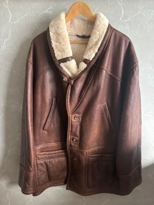 Hollies Brun skinnjacka med fårull - Snygg brun skinnjacka med bred, mjuk fårullskrage och knäppning framtill. Jackan har två stora fickor och en avslappnad passform. Perfekt för dig som vill ha en klassisk och varm look med vintagekänsla.