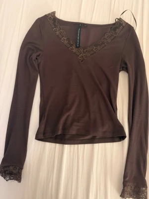 Brun långärmad topp med spets - Mörkbrun långärmad topp från The Black Label Collection i storlek XS. V-ringad med dekorativ spets runt halsringningen och vid ärmsluten. Figurnära passform och mjukt material som känns skönt mot huden. Perfekt för dig som gillar stilrena detaljer.