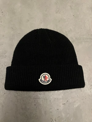 Svart Moncler mössa med logga - Snygg svart ribbstickad mössa från Moncler med uppvikt kant och klassisk Moncler-logga framtill. Perfekt accessoar för att hålla värmen och addera stilpoäng till din outfit. Tillverkad i mjukt material som sitter skönt på huvudet.