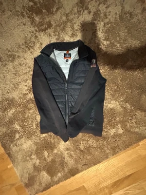 Svart Cardigan - Snygg svart cardigan med quiltad front och mjuka sidopaneler. Västen har dragkedja framtill. Perfekt för lager-på-lager och håller dig varm med lättviktigt dunfoder.