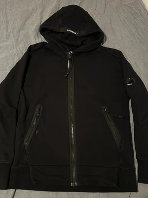 Svart hoodie från C.P. Company - Svart hoodie från C.P. Company med dragkedja framtill, två snedställda fickor med dragkedja och klassisk linsdetalj på vänster ärm. Tillverkad i mjukt material och har en stor huva. Perfekt för en stilren och avslappnad look.