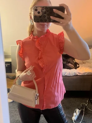 Korallrosa blus med volangdetaljer - Snygg korallrosa blus med volangkant längs knäppningen och ärmslut. Blusen är ärmlös och har en feminin, lätt figursydd passform. Perfekt att styla med jeans eller kjol för en trendig look. Materialet ser ut att vara bomull eller bomullsblandning.