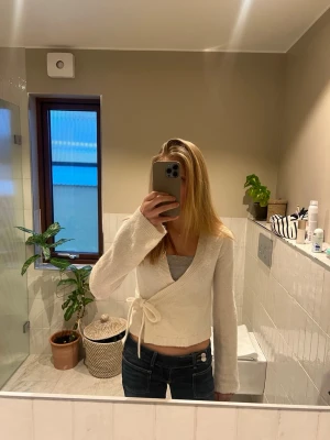 Vit stickad tröja (Gina tricot) - Säljer en vit stickad omlottopp med knytband i midjan och långa ärmar. Tröjan har en croppad passform och är gjord i ett mjukt, fluffigt material. Perfekt att styla med jeans eller kjol för en trendig look.