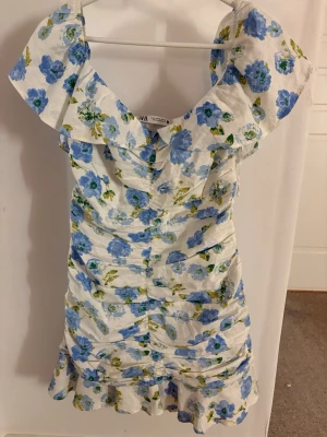 Klänning från Zara - Superfin kortklänning från Zara med blått blommönster på vit botten. Klänningen har volangdetaljer, v-ringning och breda axelband. Perfekt för sommarens alla tillfällen och ger en fräsch och romantisk look.