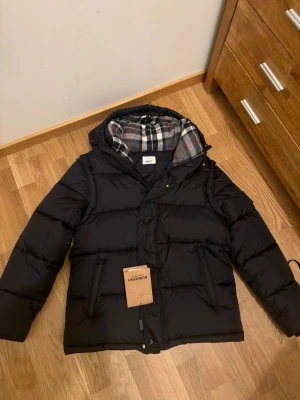 Svart pufferjacka från Burberry - Säljer en svart pufferjacka från Burberry med klassiskt rutigt foder i huvan. Jackan har huva, dragkedja och knappar framtill samt två fickor med dragkedja. På vänster ärm sitter en Burberry-patch. Perfekt för kalla dagar och riktigt snygg streetstil.