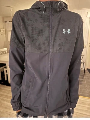 Under Amor grå vindjacka - Min brosa säljer en Under Armour,vindjacka i färgen blå/grå.Den är äkta såklart.Det här är ingen vanlig jacka utan det är en speciell Under Armour jacka.Den har camomönster överdel som är då det speciella.Materialet är lätt.Jackan har en luva,dragkedja framtill och två sidofickor och elastiska muddar.Mycket fint skick!