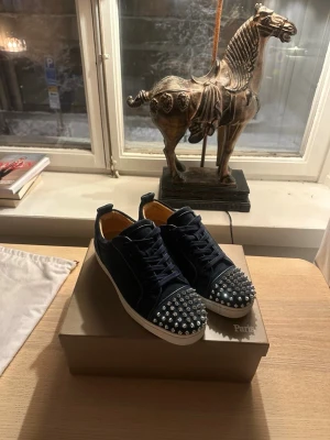 Mörkblå Christian Louboutin med gråa nitar - Säljer ett par mörkblå sneakers från Christian Louboutin i mocka med coola silverfärgade nitar på tån. Skorna har snörning, vit platt sula och beige insida i skinn. Perfekta för dig som vill sticka ut med en edgy detalj.