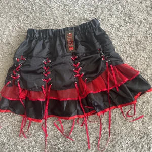 Svart och röd minikjol med snörning från Killstar - Cool svart minikjol med röda detaljer och snörning framtill och på sidorna. Kjolen har volanger och elastisk midja för extra komfort. Perfekt för dig som gillar alternativ stil och vill sticka ut.