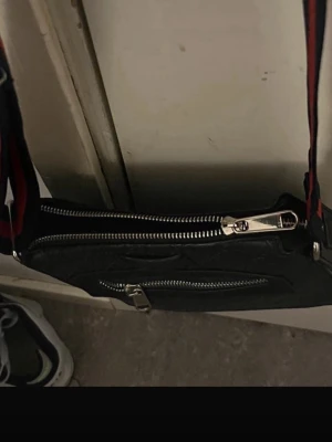 Svart axelväska med dragkedjor - Snygg svart axelväska i skinnimitation med två silverfärgade dragkedjor och justerbart axelband i rött och blått. Väskan har en stilren, fyrkantig form och passar perfekt för dig som vill ha en praktisk men trendig accessoar.