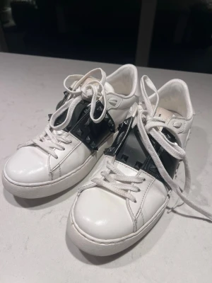 Valentino vita sneakers med nitar - Säljer ett par vita Valentino sneakers med svart band och svarta pyramidformade nitar på sidorna.  Skulle säga dne passar 38-39 också hsr det i alla skorna och dessa är inget problem då dem töjts ut lite för de har andvända men dem är i väldigt fint skick 