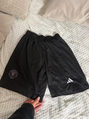 Svarta Inter Miami shorts - Säljer ett par svarta shorts med Inter Miami CF-logga på vänster ben. Shortsen har elastisk midja och är tillverkade i ett lätt och svalt syntetmaterial, perfekta för fotboll eller träning. 