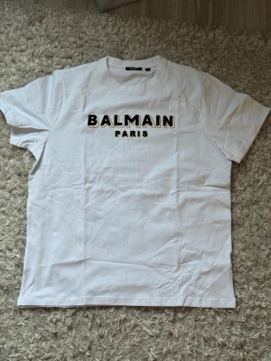 Vit Balmain Paris t-shirt - Säljer en clean vit t-shirt från Balmain Paris med svart och gul logga tryckt på bröstet. Klassisk rund hals och korta ärmar. Perfekt till jeans eller shorts för en enkel men lyxig look. Tillverkad i mjuk bomull för skön känsla.