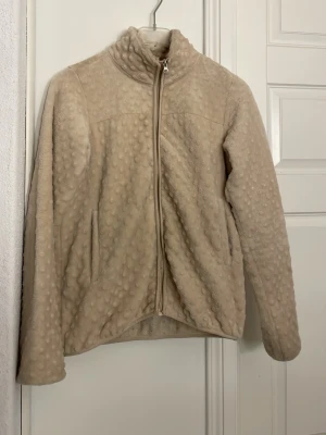 Beige teddyjacka med dragkedja - Mysig beige teddyjacka med hög krage och hel dragkedja framtill. Jackan har ett prickigt strukturmönster i materialet och två sidofickor. Perfekt för dig som vill ha en varm och trendig look under kyliga dagar. 