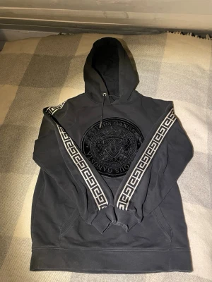 Svart hoodie från Glorious Gangsta - Snygg svart hoodie från Glorious Gangsta med stort sammetstryck på bröstet och coolt bi-motiv. Huva med dragsko och dekorativa band med grekiskt mönster längs ärmarna. Perfekt för dig som gillar streetwear och vill sticka ut lite extra. Kommer från rökfritt hem! Skickas inom 24 timmar och levereras inom 1-3 arbetsdagar 