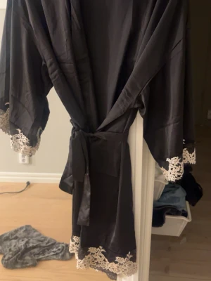 Svart kimonomorgonrock med spets - Snygg svart morgonrock i kimonomodell med breda ärmar och knytband i midjan. Dekorerad med beige spetsdetaljer vid ärmslut och nederkant som ger en lyxig känsla. Perfekt för chill dagar hemma eller när du vill känna dig extra fin.