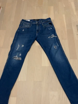 Blå Replay jeans med slitningar - Säljer ett par blå jeans från Replay med snygga slitningar och detaljer på benen. Jeansen har klassisk femficksmodell, normal passform och är tillverkade i mjukt bomullsmaterial. Perfekta för en avslappnad och trendig look.