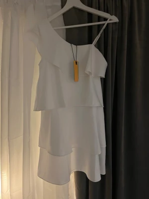 Vit omlottklänning med volanger - Supersöt vit kortklänning med lager på lager-volanger och oneshoulder-design. Klänningen har en tunn axelrem på ena sidan och bred volangkant över axeln. Perfekt för sommarens alla tillfällen!