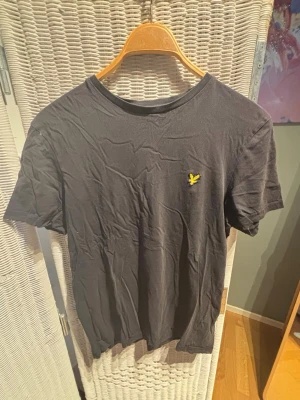 Svart t-shirt från Lyle & Scott - Klassisk svart t-shirt från Lyle & Scott med gul broderad logga på bröstet. T-shirten har rund halsringning och korta ärmar. Perfekt basplagg som funkar till det mesta och är tillverkad i mjuk bomull.