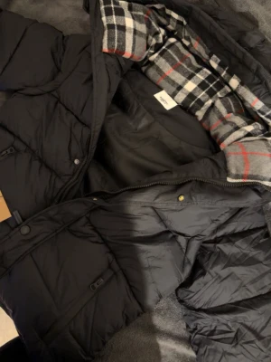 Svart pufferjacka från Burberry - Säljer en svart pufferjacka från Burberry med klassiskt rutigt innerfoder i svart, vitt och rött. Jackan har huva, dragkedja framtill och Burberry-logga på ärmen. Perfekt för kalla dagar och riktigt snygg till streetwear-stilen.