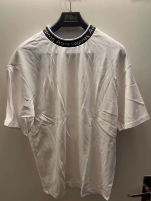 Acne Studios Logo Collar T-Shirt - Size S - Elevate your essentials with this classic Acne Studios t-shirt in white. This piece features the iconic logo-intarsia ribbed collar, making it a subtle but recognizable statement piece.  Condition: Excellent pre-owned condition. Gently used and very well cared for. No stains, holes, or discolorations.  Fit: Relaxed/Boxy fit (characteristic of Acne Studios).  Size: Small (S)  Fabric: High-quality, mid-weight organic cotton that feels premium and holds its shape.  Perfect for layering or wearing o