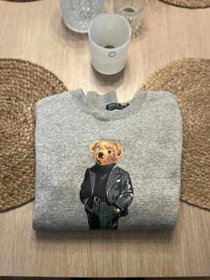 Grå sweatshirt med björn från Polo Ralph Lauren - Säljer en grå sweatshirt från Polo Ralph Lauren med den ikoniska björnen på bröstet, klädd i svart kostym och polotröja. Tröjan har rund halsringning och ribbade muddar. Perfekt för dig som gillar streetwear och klassisk stil.