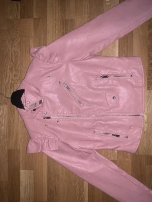 Rosa skinnjacka med rosetter - Säljer en rosa skinnjacka med snygga rosettdetaljer på axlarna. Jackan har flera dragkedjor framtill och en klassisk bikerstil. Perfekt för dig som vill sticka ut med en färgglad och trendig look.