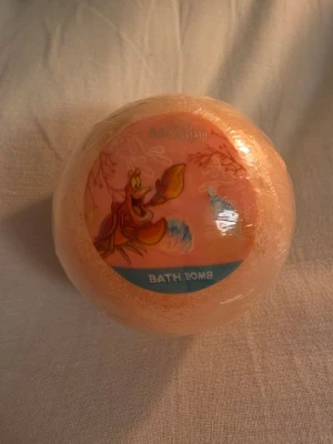 The Little Mermaid badbomb - Säljer en rund badbomb i persikofärg med motiv från The Little Mermaid och Sebastian på etiketten. Badbomben väger 100 g och är inslagen i plast. Perfekt för ett lyxigt bad med Disney-tema.