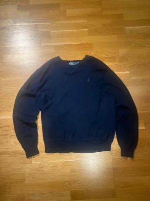 Mörkblå stickad tröja från Polo Ralph Lauren - Mörkblå stickad tröja från Polo Ralph Lauren med klassisk rund halsringning och den ikoniska gröna loggan broderad på bröstet. Tröjan har långa ärmar och ribbade muddar vid ärmslut och nederkant. Perfekt för lager på lager under kyliga dagar.