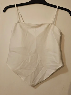 Vit linnetopp med smala axelband - Stilren vit linnetopp med smala, justerbara axelband och spetsig nederkant. Axelbanden går att ta bort så den blir off shoulder. Toppen har slät framsida och smockad baksida för skön passform. 