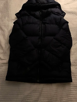 Svart dunjacka från Polo Ralph Lauren - Snygg svart dunjacka från Polo Ralph Lauren med klassisk pufferdesign och diskret logga på bröstet. Jackan har dragkedja framtill, flera fickor och en hög krage som skyddar mot kyla. Perfekt för kalla dagar och enkel att matcha med olika outfits.