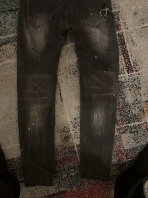 Svarta Dsquared2 ICON jeans - Säljer ett par svarta Dsquared2 ICON jeans med vit text framtill och coola slitningar samt färgstänk för en edgy look. Jeansen har smal passform och klassisk femficksdesign. Perfekta för dig som vill sticka ut med din stil.