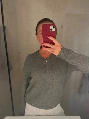 Grå stickad tröja med v-ringning - Mysig grå stickad tröja med v-ringad hals och långa ärmar. Tröjan har en klassisk och enkel design som passar perfekt till jeans eller kjol. Materialet ser ut att vara mjukt och bekvämt, perfekt för kyligare dagar.