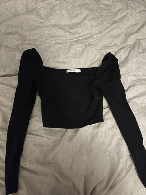 Svart croppad topp från NA-KD - Trendig svart croppad topp från NA-KD med långa puffärmar och fyrkantig urringning. Tunn och stretchig kvalitet som sitter snyggt på kroppen. Perfekt att styla med höga jeans eller kjol för en cool look.
