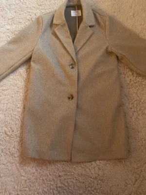 Beige ullkappa från zara 11-12 år - Stilren beige kappa i ullmix med klassisk krage och två stora knappar framtill. Långärmad modell med rak passform som ger en clean och tidlös look. Perfekt för dig som gillar minimalistisk stil.