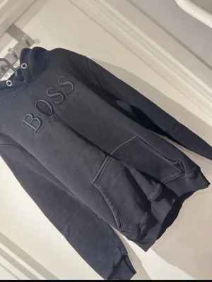 Svart hoodie från Hugo Boss - Snygg svart hoodie från Hugo Boss med stor logga broderad på bröstet. Klassisk känguruficka framtill, huva med dragsko och ribbade muddar vid ärmar och nederkant. Perfekt för en chill och stilren look. Tillverkad i mjukt sweatshirtmaterial.
