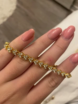 Guldigt tennisarmband med stenar - Snyggt tennisarmband i guldton med gnistrande klara stenar i varje länk. Armbandet har en klassisk och elegant design med fyrkantiga länkar och stängs med ett smidigt lås. Perfekt för dig som gillar bling och vill addera lite extra shine till din look.