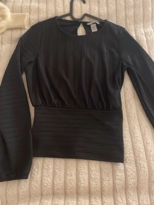 Svart långärmad topp med struktur - Snygg svart topp med lång ärm och ribbad struktur. Toppen har en bred midjedel och en liten öppning med knapp i nacken. Passar perfekt till både jeans och kjol för en trendig look.