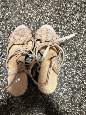 Beige kilklackade sandaler från H&M - Säljer ett par beige sandaler från H&M med flätad ovandel och knytband runt ankeln. Skorna har öppen tå och hög kilklack. Perfekta för sommaren och ger en bohemisk vibe till din outfit. Storlek 38