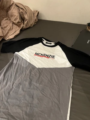 McKenzie t-shirt med kontrastärmar - Snygg t-shirt från McKenzie med svartvita kontrastärmar och grå nederdel. Tryckt logga framtill med röd detalj. Klassisk rund halsringning och sportig look, perfekt för en avslappnad stil.