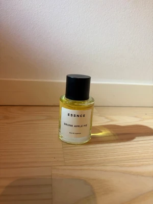 ESSNCE Drunk Apple Pie parfym - Parfymen Drunk Apple Pie från ESSNCE kommer i en rund glasflaska med svart lock. Vätskan är gul och flaskan har en stilren, minimalistisk etikett. 