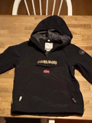 Svart vindjacka från Napapijri - Svart vindjacka från Napapijri med huva och dragkedjor på sidorna. Jackan har en stor ficka framtill med Napapijri-logga och norsk flagga. Perfekt för blåsiga dagar och har tejpade sömmar för extra skydd. Små defekter vid dragkedjan vid halsen