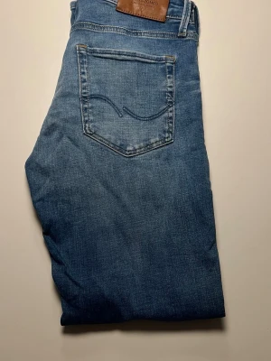Blå jeans från Jack & Jones - Snygga blå jeans från Jack & Jones med klassisk femficksdesign och ljus tvätt. Jeansen har normal passform och raka ben, samt en läderpatch med logga bak i midjan. Perfekta för en avslappnad stil. Kan gå ner i pris vid snabbaffär!!