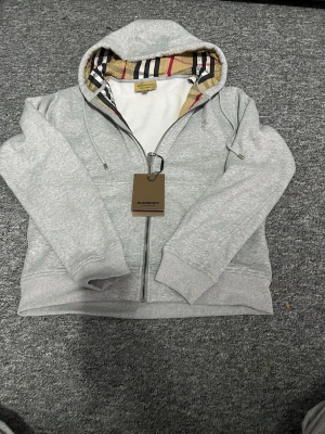 Grå hoodie från Burberry med dragkedja - Snygg grå hoodie från Burberry med dragkedja och klassiskt rutigt foder i huvan. Tillverkad i mjukt bomullsmaterial och har justerbara snören i huvan. Perfekt för dig som vill ha en stilren och exklusiv look med ikoniska Burberry-detaljer.