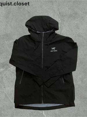 Svart vindjacka från Arc'teryx - Snygg svart vindjacka från Arc'teryx med huva och dragkedja framtill. Jackan har minimalistisk design, två sidofickor med dragkedja och logga på bröstet. Perfekt för blåsiga dagar och outdoor-äventyr. Det står storlek M men den är ganska liten i storlek så passar bättre som storlek S.