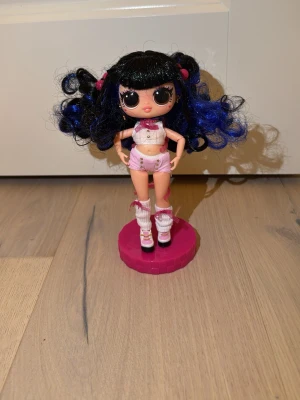 Docka med blåsvart lockigt hår - Säljer en cool docka med svart och blått lockigt hår, klädd i rosa topp och shorts, vita benvärmare och rosa skor. Dockan står på en rosa ställning och har små rosa detaljer i håret. Perfekt för dig som gillar att samla eller leka med dockor. Du får även med 1 hårborste, 1 fön, 2 par klädesplagg, 1 ryggsäck, 1 halsband samt 1 par solbrillor.