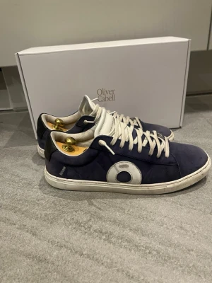 Blå mockasneakers från Oliver Cabell - Snygga sneakers från Oliver Cabell i mörkblå mocka med vita detaljer och snörning. Skorna har en vit sula och en rund tå, samt en kontrasterande vit cirkeldetalj på sidan. Perfekta för dig som gillar stilrena och moderna sneakers.  Sparsamt användna så dom är i väldigt bra skick. Box och dustbag ingår