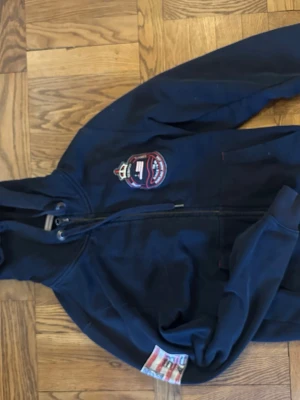 Mörkblå hoodie med dragkedja - Snygg mörkblå hoodie med dragkedja framtill och broderad patch på bröstet. Huva med snörning och ribbade muddar. Detaljer i rött, vitt och blått på insidan av ärmslutet. Perfekt för en avslappnad och sportig stil.